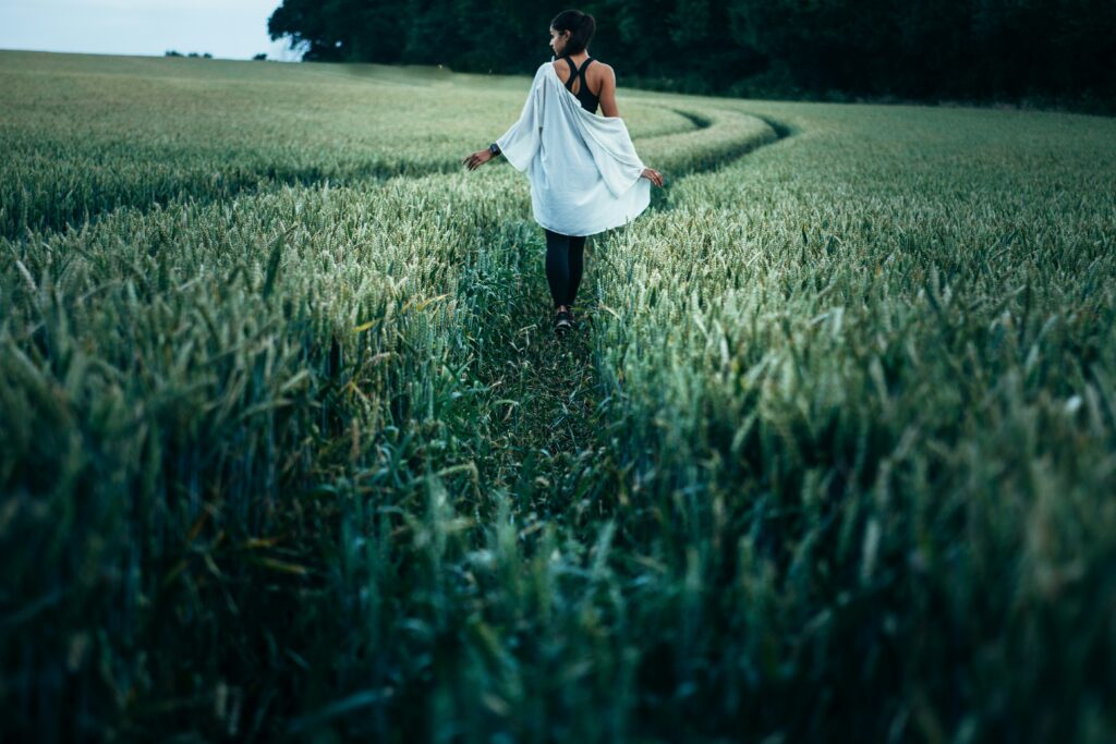 A woman walks through a field of tall grasses touching them with her hands.