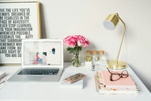 White desk with an open laptop on it in addition to journals, a lamp, and a motivational sign.